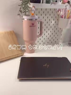 A well-lit, lifestyle shot featuring a desk scene with study essentials. Centered on a white desk, a closed laptop is positioned towards the bottom with a large pink Stanley tumbler on the left. The background includes a white pegboard with items hung.