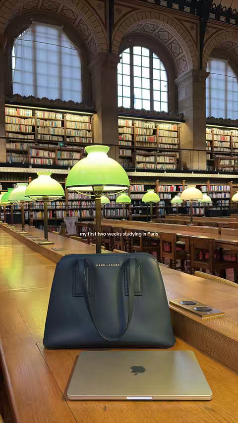 The image captures a grand library setting. The foreground features a wooden table with a navy-colored tote bag carrying the Marc Jacobs logo, a silver laptop featuring the Apple logo, and a golden phone standing on the table. Light from green lamps reflects on the dark wooden table. The background shows rows of bookshelves filled with books and elegant arching windows.