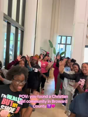 Group of young women in a brightly lit space, likely a community hall. Most are smiling and waving at the camera, and one woman in the foreground is wearing a t-shirt with text. The background includes large windows and a minimal, modern interior design.