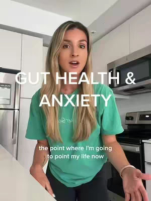 A woman stands in a kitchen setting, angled slightly towards the camera. Overlayed text reads "GUT HEALTH & ANXIETY". The woman has long blonde hair and is wearing a green t-shirt. Behind her are white cabinets and a stainless steel microwave, suggesting a domestic environment.