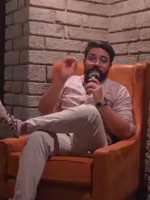 A man with a beard and glasses sits in an orange armchair holding a microphone. He's wearing a light-colored shirt and pants, and the background is a brick wall. He is gesturing with his right hand.