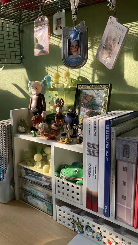 A shelf filled with various objects and figurines, set against a wall. The shelf is white and holds items of various sizes and colors. There are figurines of cartoon characters, books, and accessories like keychains. Some books are visibly labeled. The overall aesthetic is neat and organized, suggesting a personal space or collection.
