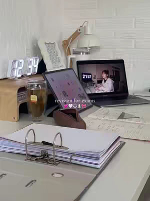 A well-lit, aesthetically organized workspace with a clear focus on studying. A laptop on the right displays a video of a person; an iPad is in the center with stickers on the screen; a glass of tea, an analog clock, and a binder are placed in front of a white brick wall in the background. The setup conveys a mood of productivity and organization.