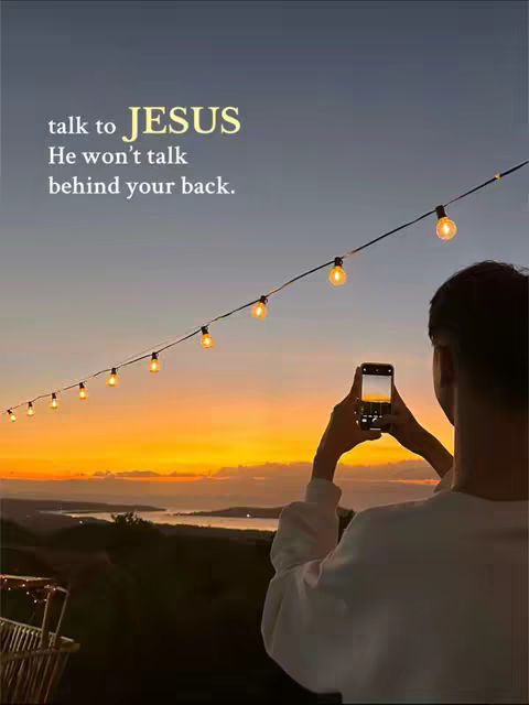 An outdoor scene at sunset with a person taking a photo of the scenery. Warm orange and yellow hues fill the sky above a dark treeline and water. Decorative string lights are strung across the top. Added overlay text provides a religious message.