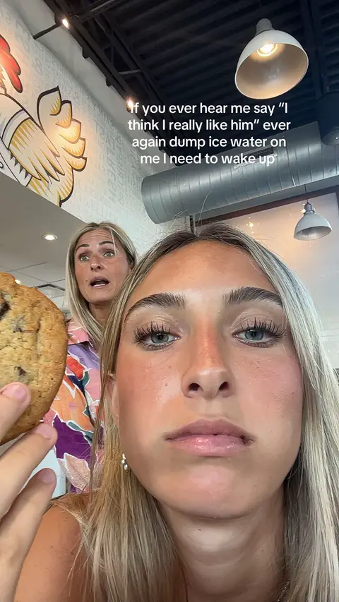 A selfie-style photo features two fair-skinned women in a restaurant setting. The woman in the foreground has blonde hair, a slightly concerned expression, and prominent eyelashes. She holds a partially visible image of a large cookie. Behind her, another woman with blonde hair looks shocked, with her mouth open, and her skin flushed. The background suggests a casual dining environment with an interior design of text and chicken imagery on the wall, and a ceiling light.