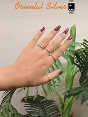 A well-lit image features a light-skinned hand with long, maroon-colored nails. The hand is adorned with multiple rings. In the background, there are green plants.