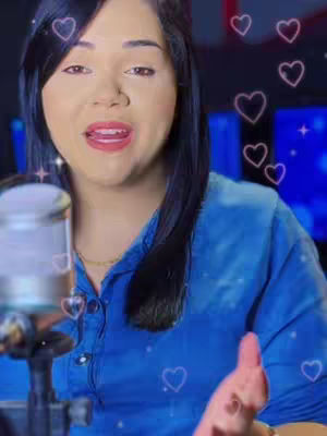 A woman with dark hair and fair skin is centered in the frame. She wears a blue button-down shirt. The shot includes a microphone and several floating pink hearts, along with a blurred background featuring studio equipment. The woman appears to be speaking or singing with an open mouth and a focused gaze, giving a sense of engagement and intimacy.