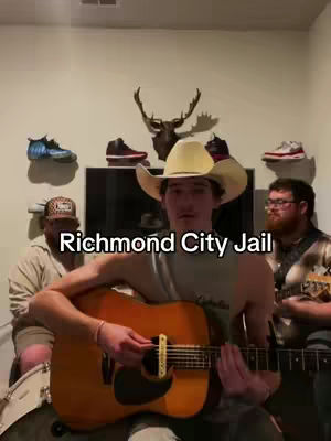 The image shows a man in a cowboy hat playing an acoustic guitar, singing with two other men playing instruments in the background. They are indoors, with the text overlayed on the image.