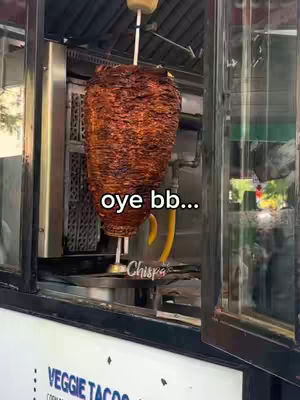 A close-up shot inside a street-food kitchen. The focus is on a large, rotating spit of cooked meat, presumably for tacos al pastor. The words 'oye bb...' are written on the image.