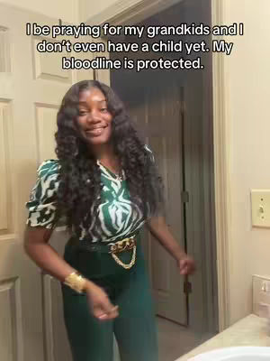 A woman with long, wavy black hair is standing in a room. She is wearing a green and white patterned top, green pants, and a gold belt, and has a gold cuff bracelet on her left wrist. The wall is visible in the background, along with a doorway and a light switch. The lighting is natural, and the overall composition is a medium shot.