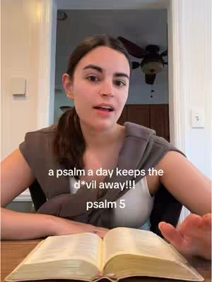 A woman sits at a table, looking toward the camera. She wears a gray hoodie draped around her shoulders. In front of her is an open book and the text overlay on the image is a religious quote.
