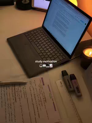 A top-down shot of a desk setup with a laptop open, displaying a document. Notes and highlighting pens are arranged around it, with a lit candle and a tablet visible behind the laptop.