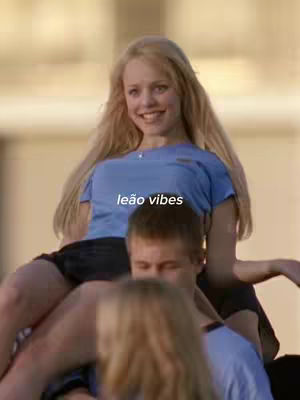 The cover image features a scene from the movie 'Mean Girls.' A blonde woman, likely portraying Regina George, is seated on the shoulders of a person with a blue shirt. The text overlay 'leão vibes' is positioned at the center of the image. The background appears to be slightly blurred, creating a focus on the subjects.