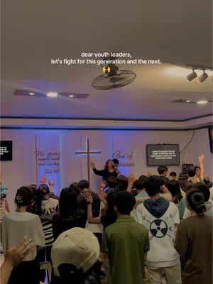 The image captures a vibrant scene at a youth leaders' camp, with a focus on a raised stage where a person is speaking, bathed in warm lighting. The audience, composed of many people, raises their hands in the air. The backdrop includes a cross and motivational texts in a church-like setting. The overall atmosphere appears spiritual and filled with communal energy.
