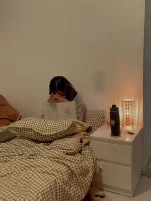 A softly lit bedroom scene features a woman sitting on a bed, leaning over, and reading a book. The bed has a checkered bedspread and pillows. A nightstand with a lamp and a water bottle sits at the side. The overall setting is calm, with warm tones and a focus on intimate activity.