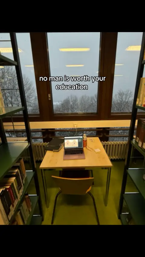A photo taken in a library study space with an emphasis on a table and window. The desk has a laptop and some items scattered on it. The window provides a view of the outside, which appears to be a cloudy day. The foreground features bookshelves on the left and right sides, typical of a library setting.