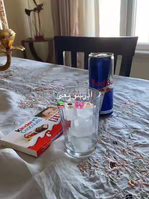 The image shows a close-up of a table set with study essentials and refreshments. A glass with ice cubes, a Red Bull can, and a Kinder Chocolate bar are arranged on a patterned tablecloth. The setting implies a study or work session.