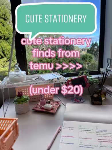 A well-lit, eye-level shot of a desk setup. The background is a window showing a green, tree-filled landscape, slightly blurred. A white desk holds stationery items including pink pen, a notebook, white candle holder, pink makeup tray, plus a peach-colored basket, and what might be a portable red game console, all near the window. Above the setup, a speech bubble graphic declares 'CUTE STATIONERY,' and further text indicates the products are from Temu and under $20.