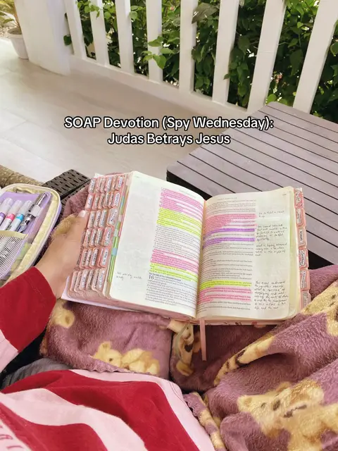 A person reading a Bible with notes and highlights, resting on a purple blanket. The background hints at an outdoor setting with a white fence and greenery.