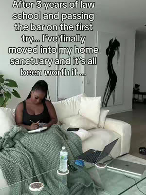 A woman sits on a cream-colored couch in a well-lit living room, writing in a notebook. She's wearing a black top and the room features a large abstract artwork, a glass coffee table with items on it, and a window with natural light.