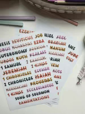 An overhead shot displays a collection of Bible verse header stickers. The stickers are colorfully designed with various names of the books of the Bible in a variety of colorful fonts. They rest on a white surface adorned with other stationary items.