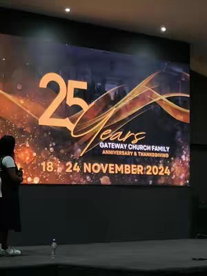 A photo shows a large digital screen displaying the words "25 Years" in large, stylized orange font, with the word "Years" in script. Behind the text, a dynamic gold and orange design sweeps across the screen. Below, it says "Gateway Church Family, Anniversary & Thanksgiving, 18. 24 November 2024." A dark-haired woman is standing in the lower left, facing away from the camera, with her back to the presentation.