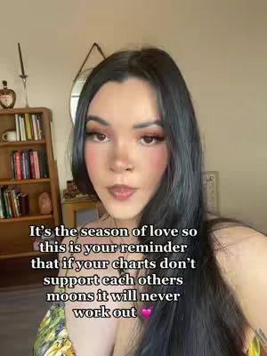 A woman with long, dark hair and makeup is centered in the frame. Text in a white font is overlaid on the screen, and the background is a room with a bookshelf, mirror, and other decor. The woman is looking forward.