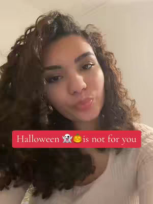 A selfie-style shot features a young woman with long, wavy, dark brown hair. She's looking directly at the camera, with a slight pout and her lips slightly parted. Overlayed on the image is a red banner with white text and emojis, conveying a direct message about Halloween.