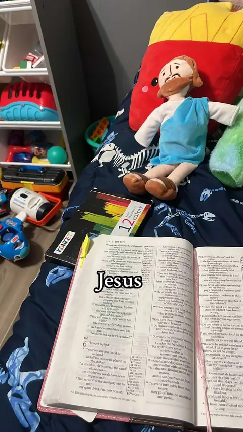 A photo taken from a slightly elevated angle shows a bed with a patterned bedsheet and various items on it. A doll of Jesus with a cloth around is sitting on the bed. A red and yellow french fries pillow is placed behind Him. Next to the doll a Bible is open with a text overlay that says 'Jesus'. Near the bottom of the image, a box of 12 colored pencils, some books, balls and toys are visible.
