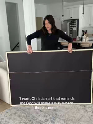 A woman in a black long-sleeved shirt stands holding a large, framed canvas with its back facing the camera. The canvas is suspended between her hands, which are at the top and bottom. The image is taken indoors with a neutral background, suggesting a home or gallery setting. In the foreground, a quote is typeset in a white, sans-serif font.