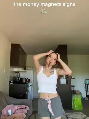 The cover image showcases a woman in a kitchen setting. She's wearing a white tank top and gray shorts with a pink drawstring. The text overlay on the image says 'the money magnets signs'. The woman is holding her hair up and has something brown in her mouth.