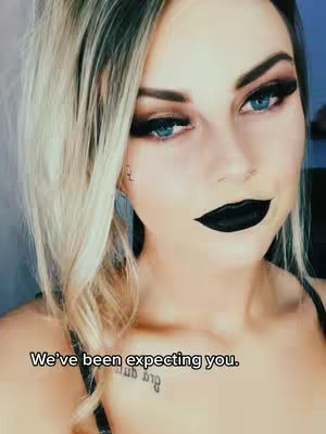 A close-up of a woman with heavy black eye makeup and black lipstick. The woman has long blonde hair with dark roots. A tattoo is visible on her upper chest. Text overlay at the bottom says, "We've been expecting you."