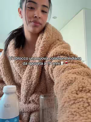 A woman, likely in her 20s, is shown close-up, wrapped in a soft, beige robe. She is styled in a ponytail. The background suggests a domestic setting, possibly a bathroom, indicating a casual, at-home vibe. Text overlayed on the image reads in Italian.