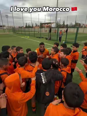 A group of children wearing orange outfits gather around a man in sunglasses, all on a green field, with the text 'I love you Morocco' on top.  A Moroccan flag is included.