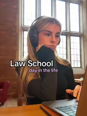 A woman is seated at a desk in a well-lit room with large windows. She is wearing a black top and headphones. In front of her is a laptop on what looks like a wooden desk, and her left arm is resting on the desk with her hand touching her face. The backdrop consists of windows and a building with red chairs.