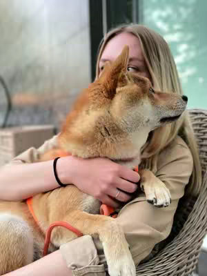 A young woman with long blonde hair is embracing a Shiba Inu dog. The dog is golden brown with a white chest and the woman is wearing a light-colored jacket. They are sitting in what appears to be an outdoor setting.