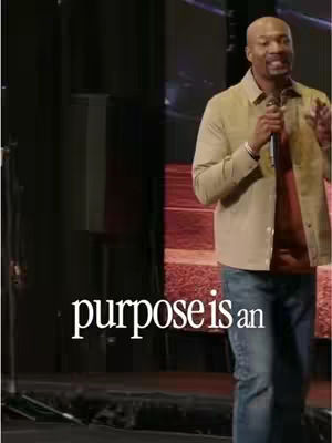 An image of a Black man on a stage, holding a microphone and speaking. Text overlaid at the bottom reads "purpose is an". The man is wearing a tan jacket, a dark red shirt, and dark blue jeans. The background is dark with stage lights.