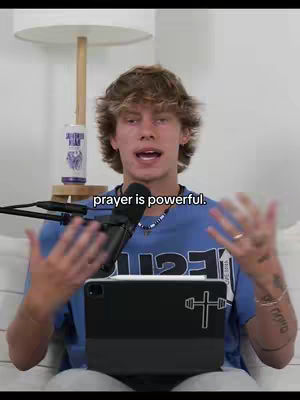 A young man with brown hair and a blue shirt is speaking into a microphone. The overlaid text 'prayer is powerful' appears in a black rectangular box at the bottom of the frame. He is sitting and has a tablet in front of him.