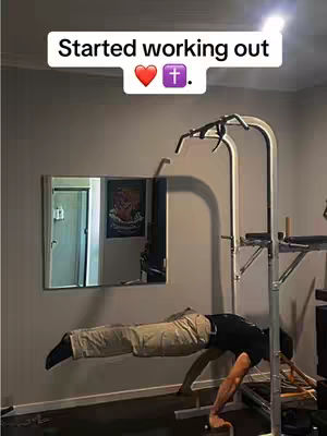 A man is doing a workout, suspended horizontally on a bar and supported by his forearms and hands on a set of parallel bars, inside a room with a mirror and a picture on the wall. The hook 'Started working out' is placed on top of the image, with a heart and a cross symbol.