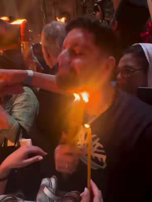 A medium shot of a man's face in a dimly lit setting. He is holding two lit candles, and the flames are touching at his neck. The background is out of focus, with other people visible, suggesting a gathering or ceremony.