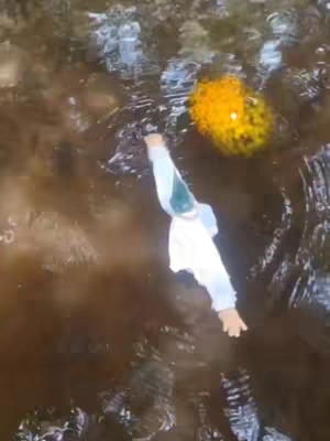 An overhead shot shows a figure, wearing a white shirt, being dragged through dark, murky water. A bright orange, almost spherical, object floats in the background. The water has distorted reflections, and the colors range from deep brown to brighter orange.