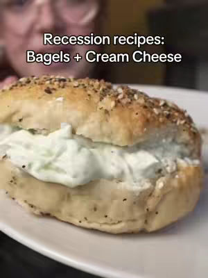 The image features a partially visible person, likely the creator, in the background with a close-up of a bagel sandwich in the foreground. The sandwich sits on a white plate, and the bagel is filled with a generous amount of cream cheese, appearing soft and white against the bagel's texture. The bagel has a dark, seedy topping. Text overlays the image, and the overall shot suggests a food-related theme, possibly a recipe or cooking demonstration.