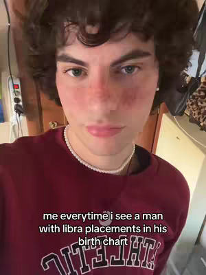 A close-up shot of a young man's face with curly brown hair. The man has pink cheeks and full lips, and is wearing a maroon sweater and a pearl necklace. Overlayed at the bottom is white text on a black background.