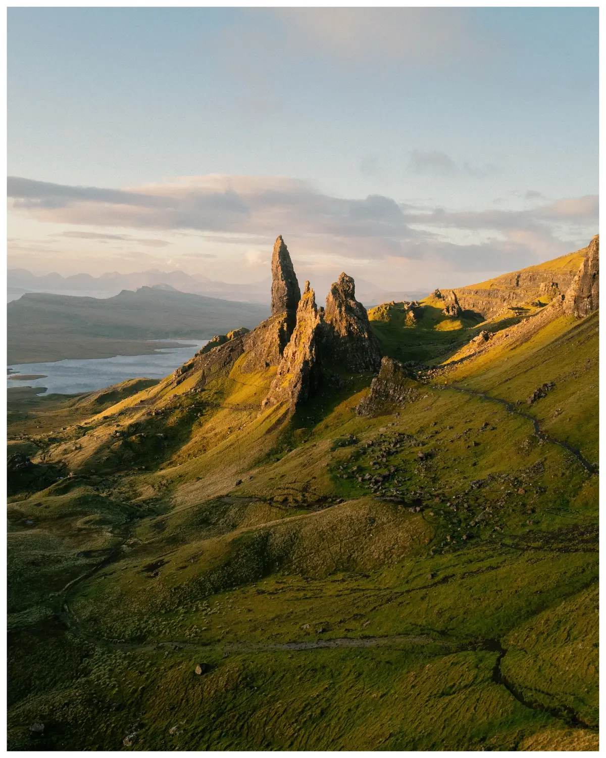 An aerial perspective of a mountainous landscape under a partly cloudy sky showcases the Isle of Skye in Scotland. Lush green hills cascade towards a body of water in the distance. In the foreground, the distinctive spires of The Storr rise towards the center, highlighted by warm sunlight. Beyond, the landscape is bathed in varying tones of green and gold, with the sky transitioning from blue to a soft, peachy hue.