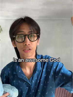 A young man with dark hair and glasses is centered in the frame. Below him is text overlayed that reads "Is an awesome God". He is wearing a blue patterned shirt. The background is plain.