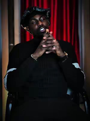 A portrait of a Black man in a dimly lit space. He is seated, wearing a black turtleneck sweater with white accents on the sleeves, and a dark cap. His hands are clasped together, with his fingers intertwined, and he appears to be gazing at the viewer. The backdrop features red curtains.
