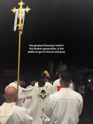 The image shows a religious procession with a tall, golden cross prominently displayed in the foreground. Priests in religious attire are walking, with the focus on a priest in the center wearing an ornate vestment. The sky is dark, suggesting the image was taken at night, and there are other people in the background, likely attending the event.