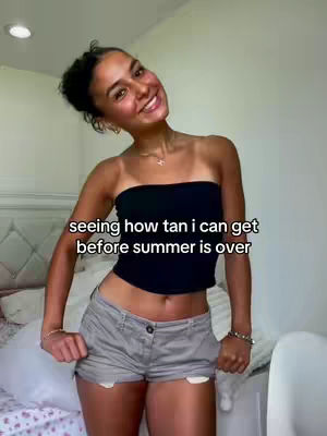 The image shows a young woman smiling at the camera. She wears a black tube top and grey shorts, revealing tanned skin with visible tan lines. The background is a room with a bed, a white headboard and white sheets. Text overlay reads: "seeing how tan i can get before summer is over".