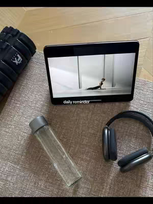 The image shows a flat lay setup on a textured taupe surface. On the left is a black foam roller. In the center, a tablet displays a video of a person doing stretches, with the text "daily reminder" at the bottom. Below the tablet is a transparent water bottle and to the right headphones resting on the surface