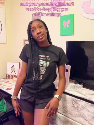 A young Black woman stands indoors, angled slightly towards the camera. She is wearing a black t-shirt with a graphic design, dark shorts, and a watch. Her long, braided hair cascades down her shoulder. The background includes a dresser, a television, and wall decorations, suggesting a college dorm or similar setting. Text overlays appear at the top.
